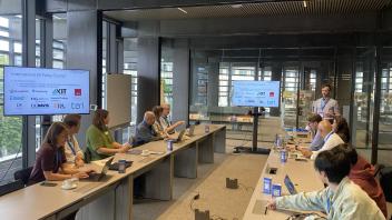 Conference room meeting around long table, participants with laptops and presentation screens