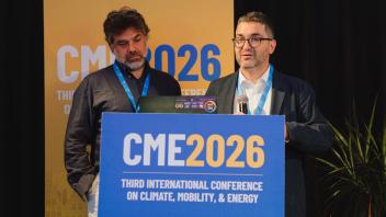 Two men at conference podium with blue 'CME2026' sign, photo