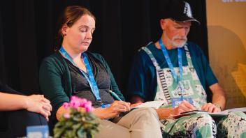 Two seated panelists with lanyards looking down on a dim stage, small potted flowers in front.