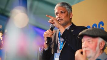 Man in suit speaking into microphone at conference; older man with cap listening (photo)