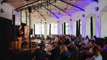Seated audience listening to a speaker at a podium in a high-ceilinged room with purple lights
