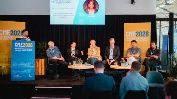 Five-person conference panel on stage with projected slide, blue banners, and seated audience