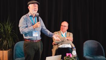 Two men on conference stage; one standing with microphone wearing hat, other seated at table.