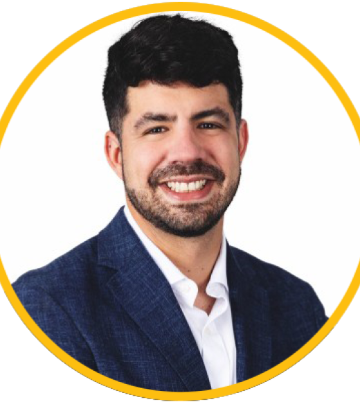 Smiling male headshot with dark hair and beard, navy blazer, yellow circular border
